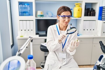 Young hispanic woman wearing scientist uniform using magnifying glass at laboratory