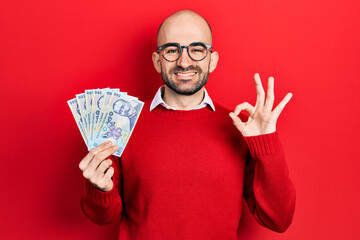 Young bald man holding 100 romanian leu banknotes doing ok sign with fingers, smiling friendly gesturing excellent symbol