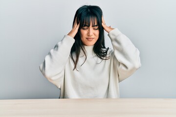 Young brunette woman with bangs wearing casual clothes sitting on the table with hand on head,...