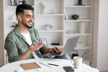 Smiling handsome millennial arabic guy teacher with beard in earphones at workplace with laptop in...