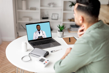 Millennial arab man checks throat, calls online to doctor on laptop screen in living room interior