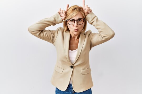 Middle age blonde business woman standing over isolated background doing funny gesture with finger over head as bull horns