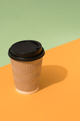 Vertical image of brown kraft paper coffee cup with plastic lid on orange and green background with copy space, mockup. Sustainable food packaging concept
