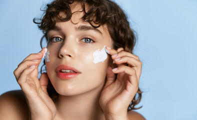 Young woman looking in mirror and applying facial cream on her cheeks, moisturizing and hydrating...