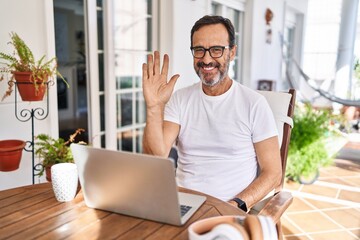 Middle age man using computer laptop at home waiving saying hello happy and smiling, friendly welcome gesture