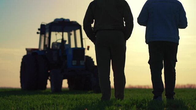 Teamwork concept. Silhouette two male farmers walking in a green field against sunset. Team farmers stand in a field on the background of agricultural machinery. Agronomists discuss harvest.