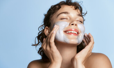 Beautiful young woman smiling, applying facial cream, scrub or cleansing product on her face with...