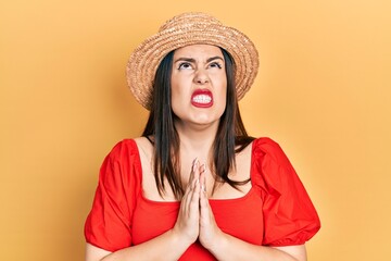 Young hispanic woman wearing summer hat begging and praying with hands together with hope expression on face very emotional and worried. begging.