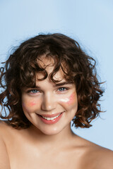 Head and shoulders. Vertical portrait of beautiful girl model with curly short hair, glitter on face and white smile, looks happy at camera, has fresh clear facial skin, blue background