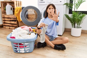 Young hispanic girl doing laundry holding socks doing stop gesture with hands palms, angry and...
