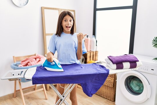 Young hispanic girl ironing clothes at laundry room pointing thumb up to the side smiling happy with open mouth