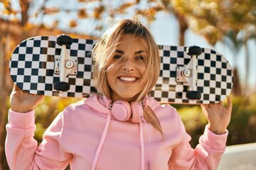 Young blonde skater girl smiling happy holding skate at the city. - obrazy, fototapety, plakaty