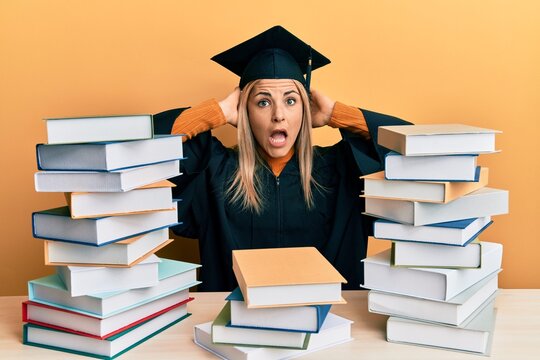 Young caucasian woman wearing graduation ceremony robe sitting on the table crazy and scared with hands on head, afraid and surprised of shock with open mouth