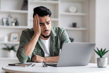 Unhappy attractive millennial muslim male with beard is sad at workplace and looks at laptop in home office - obrazy, fototapety, plakaty