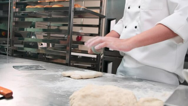 Slow-motion and close-up, chef in white cook uniforms with aprons sprinkle flour, knead pastry dough with roller, prepare bread, pies, and fresh bakery food in stainless steel kitchen of restaurant.