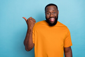 Photo of cute excited guy dressed orange t-shirt pointing thumb up empty space isolated blue color...