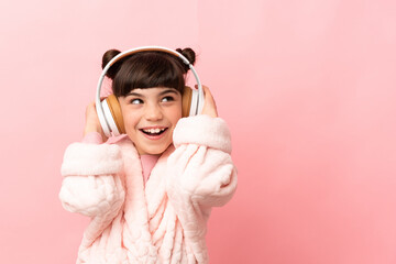 Little caucasian girl isolated on pink background in pajamas and holding a pillow and listening music