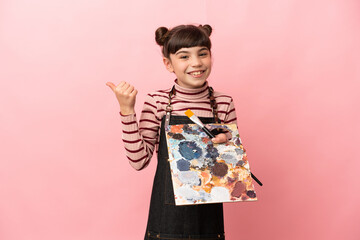Little artist girl holding a palette isolated on pink background pointing to the side to present a product