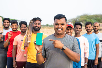 Cricket players showing green screen mobile phone by pointing finger by looking at camera at playground - concept of app promotion, advertisement and friendship