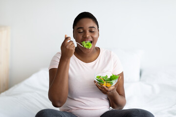 Weight loss diet concept. Overweight young black woman eating vegetable salad, sitting on bed at...