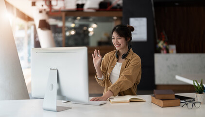 Fototapeta na wymiar Joyful young asian female in wireless headphones waving wand greeting to screen while sitting at table and having video chat with business partners using laptop against of comfortable office