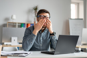 Mature male employee rubbing tired irritated eyes, working with laptop too much, exhausted from online job at office - obrazy, fototapety, plakaty