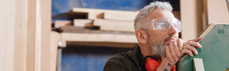 pensive grey haired furniture designer in goggles looking away in workshop, banner.