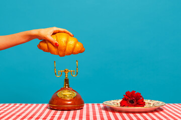 Female hand tasting crispy croissant on plaid tablecloth isolated on bright blue background. Vintage, retro style interior. Food pop art photography.