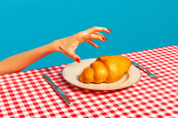 Female hand tasting crispy croissant on plaid tablecloth isolated on bright blue background. Vintage, retro style interior. Food pop art photography.