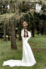 Fototapeta premium Beautiful bride in a white wedding dress and with a bouquet in her hands in a green forest