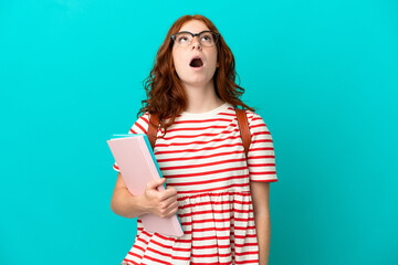 Naklejka premium Student teenager redhead girl isolated on blue background looking up and with surprised expression