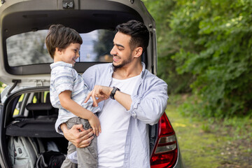 Happy arab man holding son in arms and playing with him near car with open trunk outdoor in summer