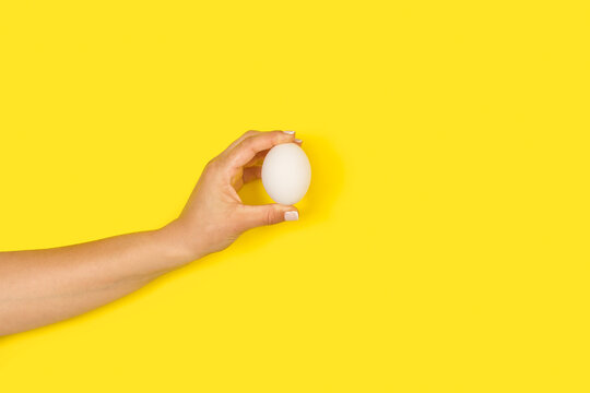 Mano de mujer sosteniendo un huevo blanco sobre un fondo amarillo liso y aislado. Vista de frente y de cerca. Copy space