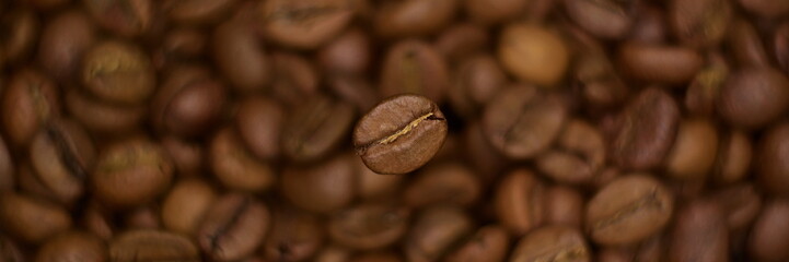 Wide angle panorama – top view natural roasted coffee bean close up hovering in frozen motion on blurry background of coffee color of heap beans. Selective focus, bokeh effect