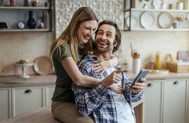 Cheerful millennial caucasian wife hugging husband with stubble and looking at smartphone in modern...