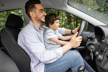 Happy millennial dad teaching little boy with steering wheel to drive car in summer, outdoor, profile