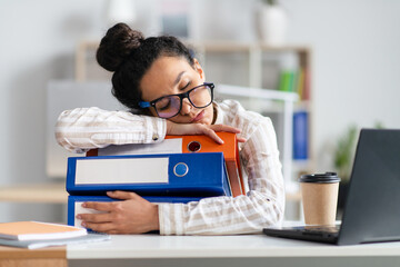 Tired businesswoman sleeping on stack of folders at her desk in front of laptop, feeling exhausted at office - obrazy, fototapety, plakaty