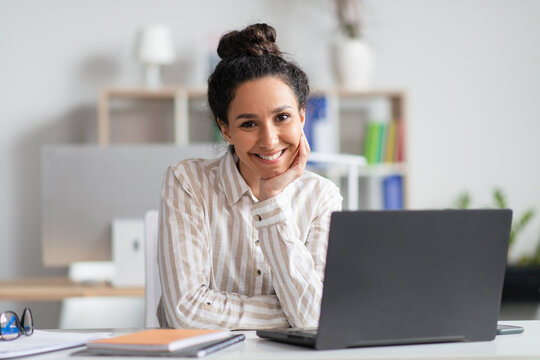 Portrait of happy businesswoman sitting at her workplace in front of laptop and smiling at camera at modern office