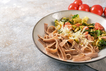 Healthy dish from whole grain tagliatelle pasta with vegetables like broccoli, tomatoes, and spinach, served with parmesan in a plate on a gray rustic stone background, copy space, selected focus
