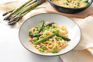 Spaghetti carbonara variation with green asparagus served in a plate, ingredients and napkin on a white table, copy space, selected focus