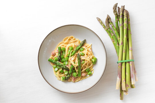 Green asparagus with spaghetti, bacon and carbonara sauce on a plate beside a bundle of the raw vegetable on a white painted wooden table, copy space, high angle view from above