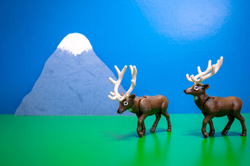 Toy deer walk along a grassy field against the backdrop of a high mountain
