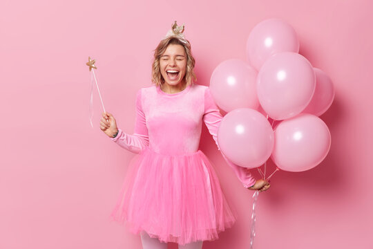 Joyful upbeat woman laughs happil has overjoyed expression holds bunch of inflated balloons and magic wand celebrates university leaving isolated over pink background. Festive occasion concept