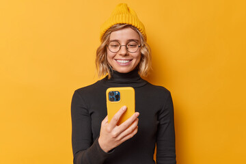 Happy positive beautiful woman with fair hair holds smartphone broses social media content connected to internet downloads new application on new gadget wears yellow hat and black turtleneck