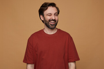 indoor portrait of bearded male posing over beige background wears red t-shirt smiles and looks into camera with positive facial expression