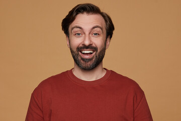 indoor portrait of bearded male posing over beige background wears red t-shirt smiles and looks into camera with positive surprised facial expression