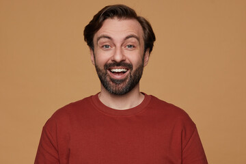 Fototapeta premium indoor portrait of bearded male posing over beige background wears red t-shirt smiles and looks into camera with positive surprised facial expression