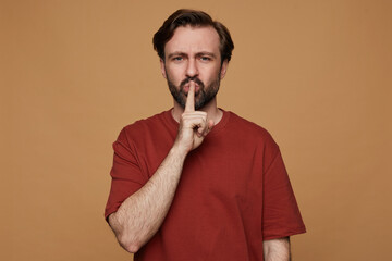 indoor portrait of bearded male posing over beige background wears red t-shirt shows silence gesture with angry, unhappy facial expression