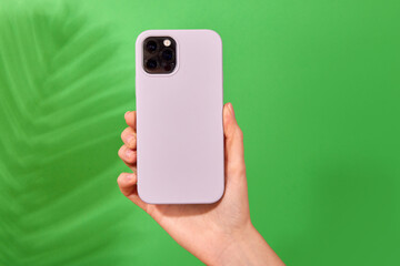 Female hand holding white smartphone in soft silicone cover back view . Phone case mock up isolated on green background shadow from a plant - obrazy, fototapety, plakaty