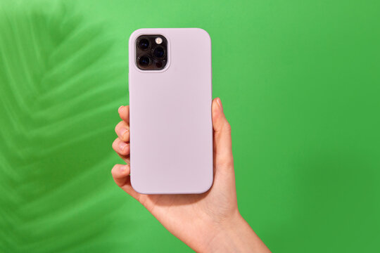 Female hand holding white smartphone in soft silicone cover back view . Phone case mock up isolated on green background shadow from a plant